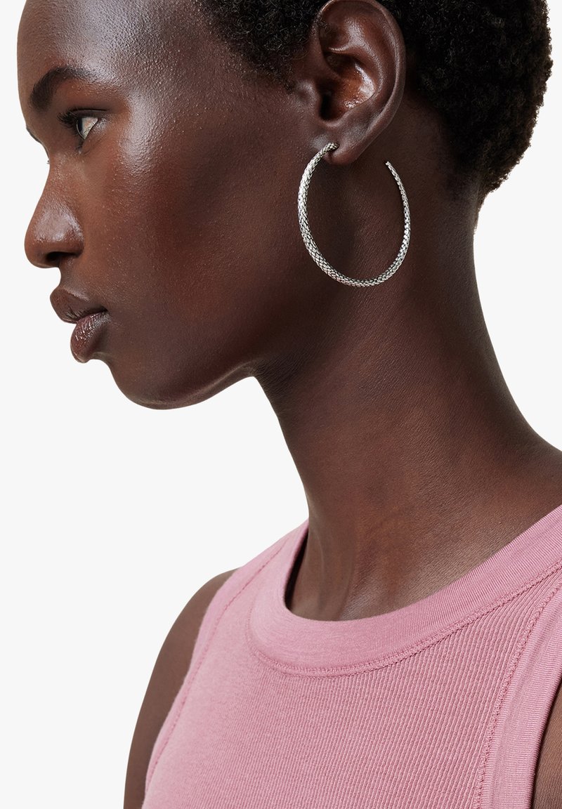 Pendientes de aro plateados con un patrón texturizado, exhibidos en un modelo con cabello corto y oscuro, que lleva una blusa sin mangas rosa.