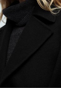 Manteau en laine noire avec un col cranté superposé à un col roulé gris foncé. Texture lisse, design minimaliste, sans quincaillerie visible.