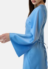Robe en satin bleu à la silhouette ajustée, avec de longues manches évasées et un détail de lacet dans le dos. Texture lisse et couleur vive.