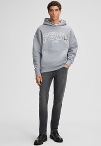 Giovane uomo che indossa un felpa FSWL grigio chiaro con ricamo, jeans grigio scuro e stivali in pelle nera, in piedi con una mano in tasca.
