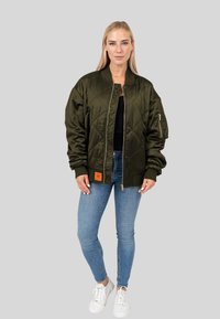 Giacca bomber verde oliva con design trapuntato, chiusura con zip, polsini a coste e un'etichetta arancione sull'orlo, abbinata a jeans blu e sneakers bianche.