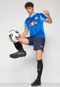 Maglia da calcio blu con il logo della Red Bull, pantaloni neri e scarpe da calcio nere. Il giocatore sta palleggiando un pallone da calcio bianco e nero.