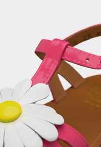 Ροζ λουρί σανδαλιού με γυαλιστερό φινίρισμα, που διαθέτει μεγάλη διακόσμηση λουλουδιού με λευκά και κίτρινα πέταλα, και καφέ πέλμα ορατό στο φόντο.