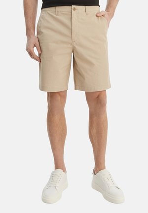 Pantalones cortos beige hasta la rodilla usados por una persona de pie con una mano en el bolsillo, combinados con zapatillas blancas sobre un fondo liso.