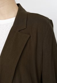 Gros plan sur une personne portant un blazer marron foncé par-dessus une chemise blanche texturée, mettant en évidence le col et l'épaule du blazer.