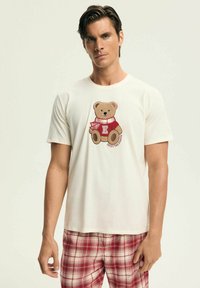 Kremowa koszulka z grafiką misia w czerwonym szaliku i swetrze z logo "E". Stylizowana z czerwonymi spodniami piżamowymi w kratę.