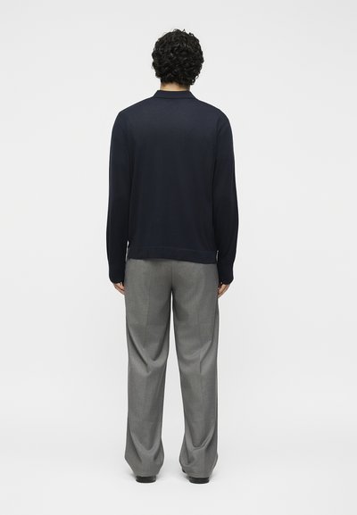 Άντρας με σγουρά μαλλιά, που φοράει μαύρο μακρυμάνικο τοπ και άνετο γκρι παντελόνι, στέκεται με την πλάτη σε ένα απλό λευκό φόντο.