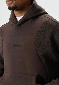 Bruine hoodie met een gestructureerde, patroonmouw, voorzien van een buidelzak aan de voorkant en bedrukte tekst met de woorden "BYE BYE LOSER." Gladde stofafwerking.