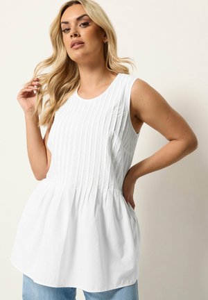 Blondynka o falowanych włosach, ubrana w białą plisowaną bluzkę bez rękawów i jasnoniebieskie dżinsy, stojąca na jednolitym tle.