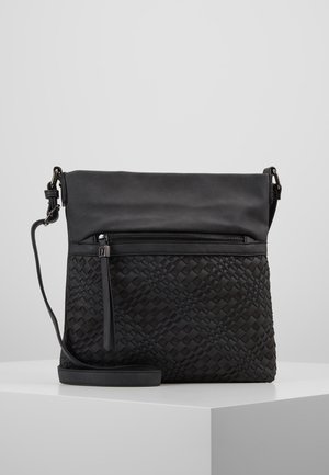 Musta olkakassi, jossa on etupuolella kudottu kuvio, taitettava yläosa, etutasku vetoketjulla ja säädettävä olkahihna, valkoisella pinnalla.