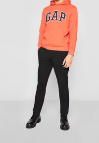 Persona vestida con una sudadera naranja brillante de GAP, pantalones negros y botas de cordones negras, de pie contra un fondo gris liso.