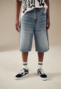 Hellblaue Jeansshorts mit weiter Passform und ausgefransten Säumen, kombiniert mit schwarzen Sneakern und weißen Socken mit schwarzen Streifen.
