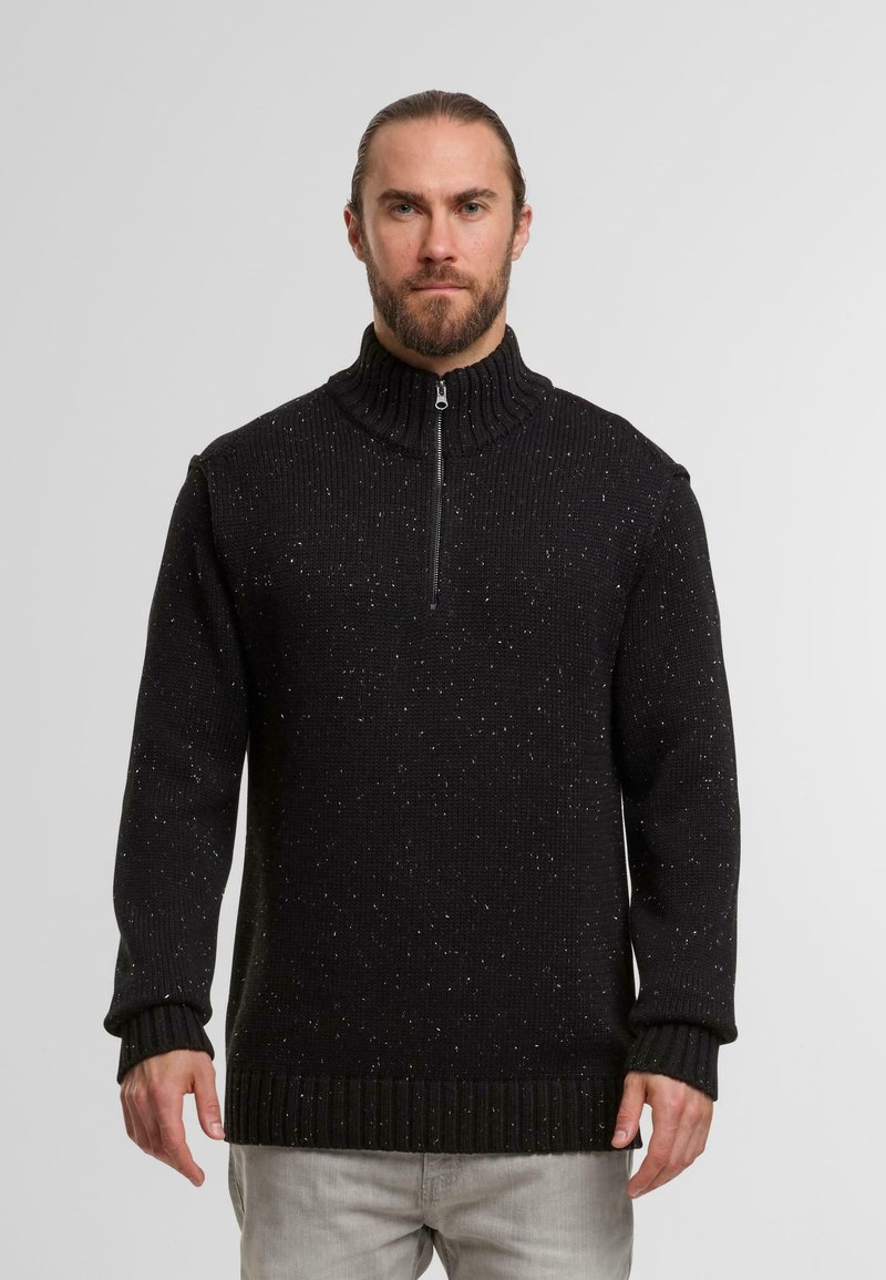 Schwarzer Strickpullover mit hohem Kragen und halbem Reißverschluss, der verstreute weiße Sprenkel aufweist. Gerippte Bündchen und unterer Saum sorgen für Struktur.