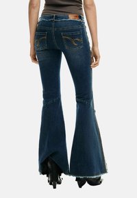 Püksid keskmise pesuga lehvikukujuliste denimist, kaunistatud tikanditega taskutel, sasitud äärega ja kitseneva vöökohaga.