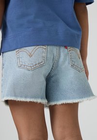 Jasnoniebieskie szorty jeansowe z postrzępionym dołem, wyposażone w klasyczne tylne kieszenie i kontrastowe pomarańczowe przeszycia, noszone z niebieskim topem.