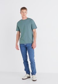 Camiseta de algodón verde con cuello redondo, combinada con jeans azules y zapatillas blancas con acentos marrones. Diseño simple y casual.