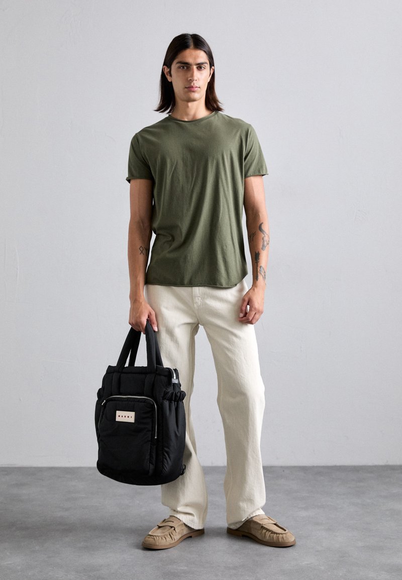 Olive green cotton t-shirt, cream wide-leg pants, tan shoes, holding a black padded bag with short handles and front zip pocket.