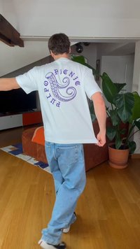 Άνδρας με ανοιχτό μπλε τζιν και λευκό μπλουζάκι με μοβ σχέδιο πλοκάμι χταποδιού και κείμενο στην πλάτη, σε σαλόνι με φυτά.