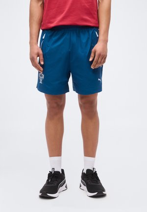 Person wearing blue athletic shorts with white logos, a red shirt, white socks, and black running shoes standing against a plain background.