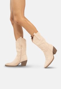 Beżowe botki z sztucznego zamszu, kostkowe, z zamkiem błyskawicznym po boku, spiczastym noskiem i teksturowanym słupkowym obcasem. Zaprojektowane z akcentami w stylu zachodnim.