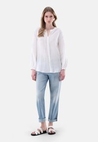 Blusa bianca di lino con dettaglio a fiocco, maniche a palloncino, abbinata a jeans azzurri arrotolati e sandali neri con cinturini bianchi.