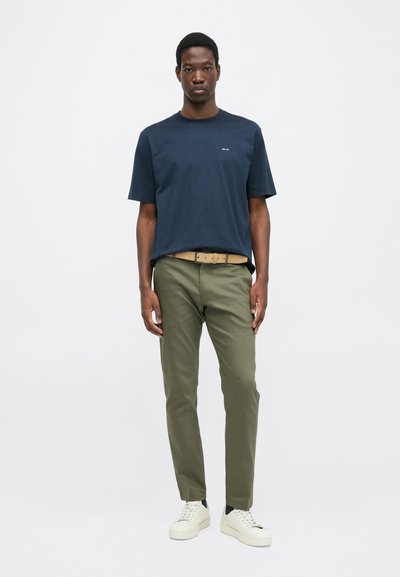 Ein Mann trägt ein marineblaues T-Shirt, olivgrüne Hose mit einem beigen Gürtel und weiße Sneakers und steht vor einem einfarbigen hellen Hintergrund.