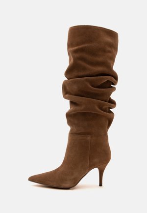 Botte marron en daim décontractée montant jusqu’au genou, avec un bout pointu et un talon stiletto fin de hauteur moyenne sur fond blanc.