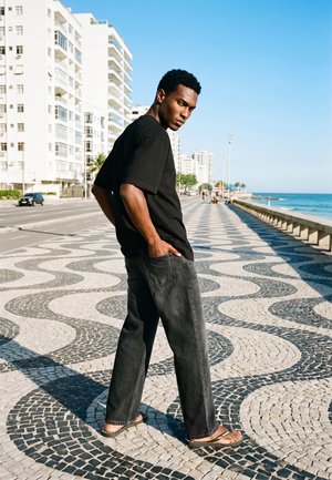 Un homme portant une chemise noire, un jean foncé et des sandales pose sur une promenade en bord de mer au motif ondulé, avec des bâtiments blancs et l’océan derrière lui.