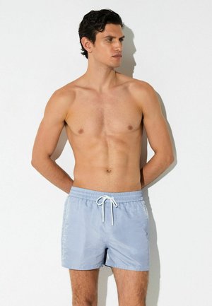 Särgi eemaldanud mees tumedate juustega, kandes helerohelisi ujumispükse valge paelaga, seisab valge tausta ees, käed selja taga.