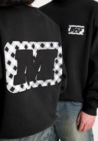 Schwarzer Sweatshirt mit großem, texturiertem "M"-Logo auf einem schwarz-weißen Karomuster. Glattes Material mit gerippten Bündchen.