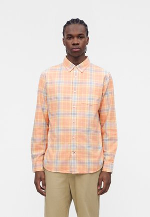 Νέος άνδρας με ροδακινί και μπλε καρό πουκάμισο με μακριά μανίκια και μπεζ παντελόνι, στέκεται μπροστά σε απλό λευκό φόντο.