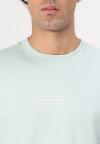 Lys grøn bomulds T-shirt med rund hals og logo på brystet. Blød tekstur, minimalistisk design, med hvid trykt branding.