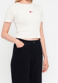 Femme portant un t-shirt court blanc avec deux petits dessins de cœurs et un pantalon noir taille haute, une main dans la poche.