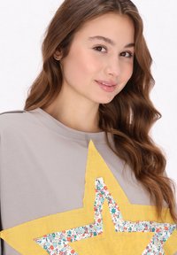Grå sweatshirt med et stort, gult stjerneapplikation med et blomsterstofmønster. Stoffet har en glat tekstur og en afslappet pasform.