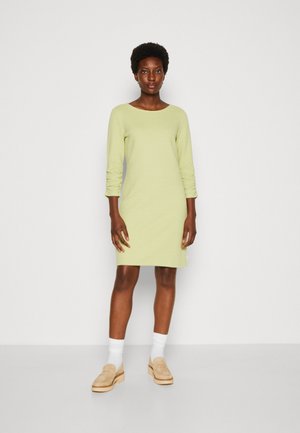 Ανοιχτό πράσινο φόρεμα με μακριά μανίκια και στρογγυλή λαιμόκοψη, φτιαγμένο από υφασμάτινη ύλη, συνδυασμένο με μπεζ λοφάκια και λευκές κάλτσες. Απλός, ίσιος σχεδιασμός.