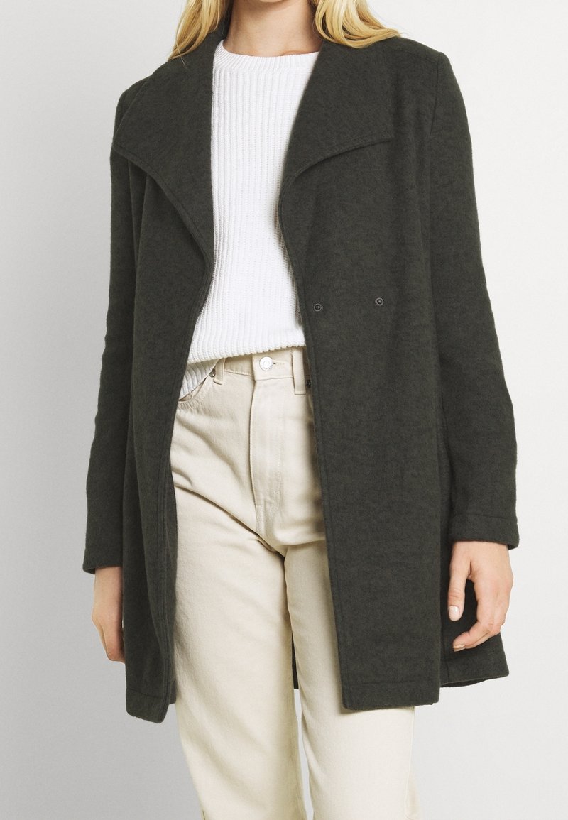 Cappotto in misto lana verde scuro con ampio revers, design doppiopetto e vestibilità rilassata. Indossato sopra un maglione bianco a costine e pantaloni beige chiaro.