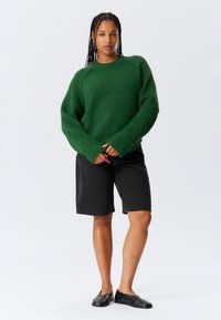 Persoon met gevlochten haar die een groene gebreide trui, zwarte knielange shorts en zwarte instapschoenen draagt, staand tegen een effen achtergrond.