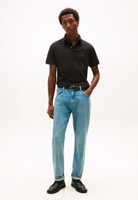 Uomo in piedi con una mano in tasca, indossa una polo nera, jeans azzurri, cintura nera e scarpe nere su uno sfondo bianco semplice.