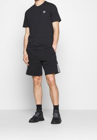 Muž ve černém tričku Adidas, černých šortkách s bílými pruhy, černých ponožkách a černých teniskách, stojící na bílém pozadí.