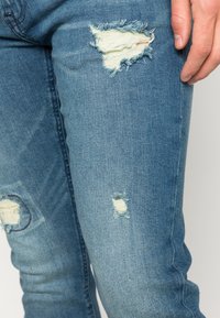 Vaqueros de mezclilla azul claro con desgarrones deshilachados en el muslo y la rodilla. La tela tiene una textura suave con un ligero estiramiento.