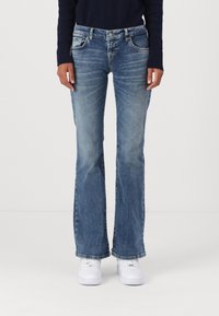 Blaue Schlagjeans aus Denim, ausgestattet mit Fronttaschen, einem ausgewaschenen Look und einem Knopfverschluss am Bund. Dazu werden weiße Sneaker getragen.