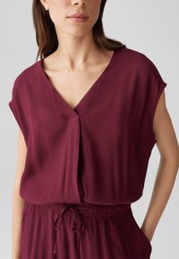 Bordeauxfarbene Bluse mit V-Ausschnitt und kurzen Ärmeln. Aus weichem Stoff gefertigt, verfügt sie über einen Vorderfalten und ein Bindedetail in der Taille.