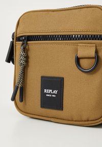 Beige canvas crossbody taske med sort lynlås, tekstureret overflade og rektangulær form. Har mærkets patch og dekorativ lynlås træk.