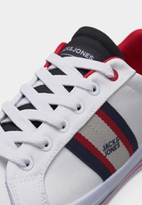 Άσπρο καμβαδένιο αθλητικό παπούτσι με ναυτικού μπλε και κόκκινες ρίγες, λευκά κορδόνια, κόκκινες οπές και μαύρη γλώσσα με το λογότυπο "Jack & Jones".