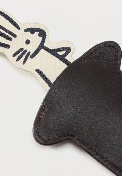 Objet en cuir noir en forme de tête de chat avec des bords cousus à côté d'un patch en tissu crème présentant un dessin au trait noir simple d'un chat.
