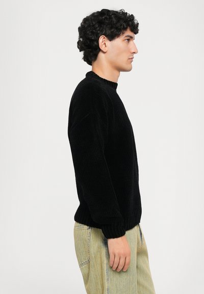 Black ribbed knit sweater with a rounded neckline, relaxed fit, and long sleeves, paired with light-colored, loose-fitting pants.