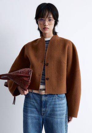 Braune Wolle Kurzjacke mit rundem Ausschnitt und zwei Knöpfen, kombiniert mit einem gestreiften Hemd und blauen Jeans; hält eine rote geflochtene Tasche.