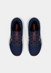 Μπλε αθλητικά παπούτσια με υφασμάτινη επιφάνεια υφής, πορτοκαλί λεπτομέρειες και λευκή σόλα. Τα κορδόνια είναι σκούρα και περνούν μέσα από τέσσερις θηλιές σε κάθε πλευρά.