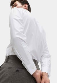 Bílé dlouhé button-up košile s detaily na manžetách, spárované se šedými texturovanými kalhotami s kapsou na zadní straně a knoflíkem.
