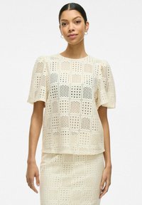 Top color crema con mangas cortas abullonadas, que presenta un patrón de rejilla con cortes. Textura suave y ajuste relajado, combinada con una falda a juego.